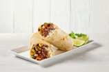 Two burritos filled with ingredients like ground meat, black beans, corn, diced peppers, and cheese are placed on a white rectangular plate. Lime wedges and cilantro garnish the display. The background is a light wooden texture.