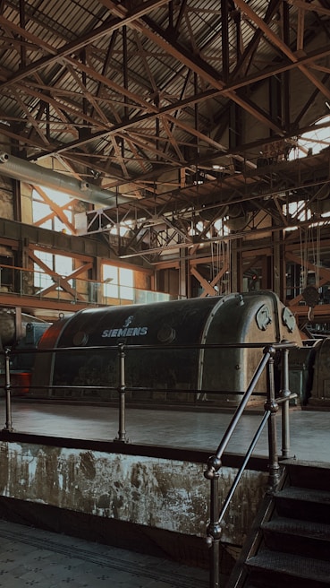 An industrial interior features a large, vintage Siemens machine encased in a metallic structure. The roof is supported by a complex grid of metal beams, allowing a soft, diffused light to filter through its large windows. The overall atmosphere is characterized by aged industrial aesthetics, with rust and wear visible on the surfaces.