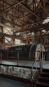 An industrial interior features a large, vintage Siemens machine encased in a metallic structure. The roof is supported by a complex grid of metal beams, allowing a soft, diffused light to filter through its large windows. The overall atmosphere is characterized by aged industrial aesthetics, with rust and wear visible on the surfaces.