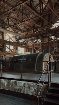 An industrial interior features a large, vintage Siemens machine encased in a metallic structure. The roof is supported by a complex grid of metal beams, allowing a soft, diffused light to filter through its large windows. The overall atmosphere is characterized by aged industrial aesthetics, with rust and wear visible on the surfaces.