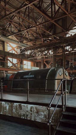 An industrial interior features a large, vintage Siemens machine encased in a metallic structure. The roof is supported by a complex grid of metal beams, allowing a soft, diffused light to filter through its large windows. The overall atmosphere is characterized by aged industrial aesthetics, with rust and wear visible on the surfaces.