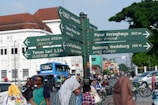 A busy city street scene with a directional signpost indicating various local attractions and facilities. The signpost lists locations and distances in both Latin and Javanese script. In the background, multiple vehicles, including cars and motorcycles, are on the road, and several pedestrians are walking. The architecture includes a mixture of colonial-style and modern buildings. There are trees lining the street, adding greenery to the urban environment.