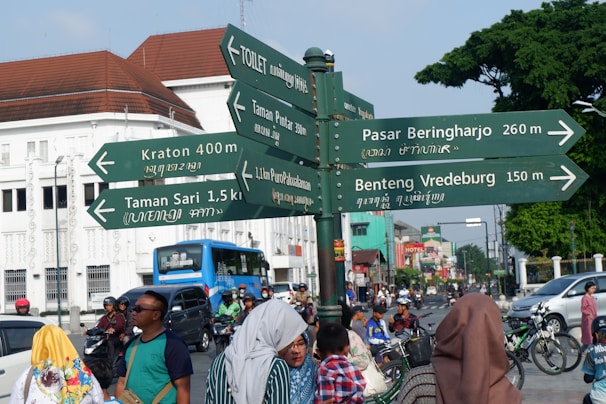 A busy city street scene with a directional signpost indicating various local attractions and facilities. The signpost lists locations and distances in both Latin and Javanese script. In the background, multiple vehicles, including cars and motorcycles, are on the road, and several pedestrians are walking. The architecture includes a mixture of colonial-style and modern buildings. There are trees lining the street, adding greenery to the urban environment.