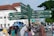 A busy city street scene with a directional signpost indicating various local attractions and facilities. The signpost lists locations and distances in both Latin and Javanese script. In the background, multiple vehicles, including cars and motorcycles, are on the road, and several pedestrians are walking. The architecture includes a mixture of colonial-style and modern buildings. There are trees lining the street, adding greenery to the urban environment.