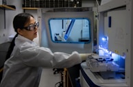 A person wearing safety goggles and a lab coat operates a scientific instrument in a laboratory setting. The instrument appears to be a complex, high-tech machine, possibly used for analysis or experimentation. The environment is clean and well-organized, with other lab equipment visible in the background.