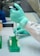 A person wearing a green glove is using a pipette to transfer liquid into a test tube held in a green rack. The workspace is clean and organized, typical of a laboratory environment.