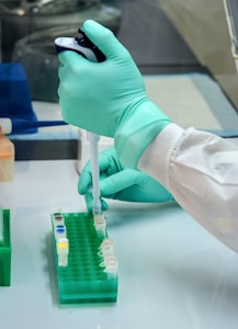 A person wearing a green glove is using a pipette to transfer liquid into a test tube held in a green rack. The workspace is clean and organized, typical of a laboratory environment.