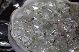 A close-up of a bowl filled with clear, sparkling crystal-like stones with metallic hooks interspersed among them. The bowl is white with a gold rim, adding a touch of elegance.
