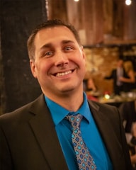 A man wearing a dark suit, teal shirt, and patterned tie is smiling at the camera. The background features a dimly lit room with a brick wall and blurred figures engaged in conversation.