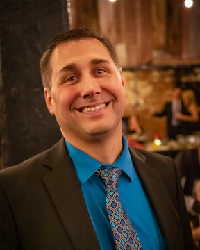 A man wearing a dark suit, teal shirt, and patterned tie is smiling at the camera. The background features a dimly lit room with a brick wall and blurred figures engaged in conversation.