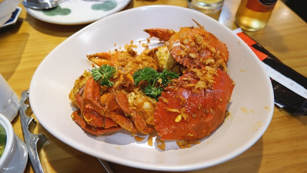 A large plate of cooked crabs garnished with herbs and crispy fried bits is placed on a wooden table. The crabs are bright red-orange, suggesting they've been cooked with spices. Part of a dining setup, including utensils, is visible in the background.
