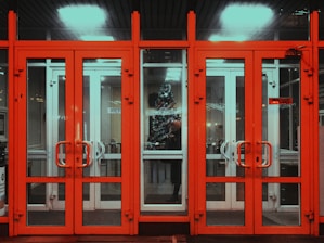 A set of bright red-framed glass doors revealing a hallway interior. Through the glass, a decorated Christmas tree is visible, adding a festive atmosphere. The lighting inside contrasts with the dark and vibrant colors of the frame.