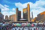 A large crowd gathers at an outdoor public space featuring the iconic curved twin-towered building complex of Toronto City Hall. The scene is vibrant with the Toronto sign prominently displayed, surrounded by people skating and socializing. Stalls with white tents add a festive ambiance. The sky is bright with fluffy clouds, and surrounding skyscrapers add to the urban landscape.