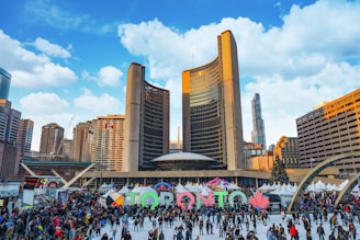A large crowd gathers at an outdoor public space featuring the iconic curved twin-towered building complex of Toronto City Hall. The scene is vibrant with the Toronto sign prominently displayed, surrounded by people skating and socializing. Stalls with white tents add a festive ambiance. The sky is bright with fluffy clouds, and surrounding skyscrapers add to the urban landscape.