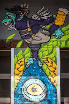 A colorful mural of a fantastical creature with a turquoise face, sharp teeth, and wings. The creature is wearing a cap and overalls, holding a mug of beer. The lower part of the mural features a large blue eye with yellow details surrounded by green and yellow foliage.