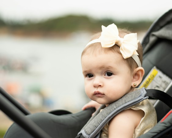 Baby in a cream hair bow sitting in an infant car seat, the harness shoulder pad visible beside her