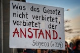 A person holds a sign with a quote in German that reads 'Was das Gesetz nicht verbietet, verbietet der Anstand. Seneca (v.334).' The text is prominently displayed, using black and red letters on a white background, and stands out against the blurred backdrop of a crowd and a partly cloudy sky.