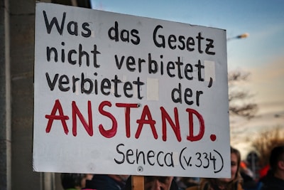 A person holds a sign with a quote in German that reads 'Was das Gesetz nicht verbietet, verbietet der Anstand. Seneca (v.334).' The text is prominently displayed, using black and red letters on a white background, and stands out against the blurred backdrop of a crowd and a partly cloudy sky.