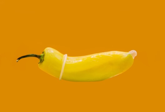 A yellow pepper is wrapped in a transparent condom against a solid orange background. The image is simple, highlighting the pepper and the unusual use of the condom.