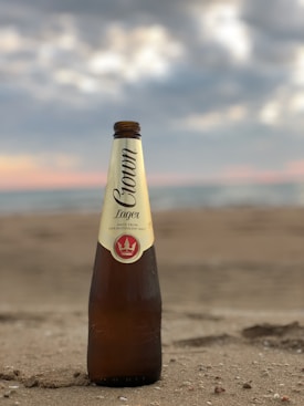 A brown glass bottle of Crown Lager stands upright on the sandy beach, with blurred ocean waves and a cloudy sky in the background.