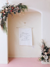 A wall decoration features a hanging white cloth inscribed with cursive text: 'Always leave room for magic and dessert'. Adorning the top of the wall arch is a lush arrangement of flowers and greenery, while a smaller floral arrangement sits on the floor to the right. The wall and arch are light cream-colored, and the floor is a soft pink.