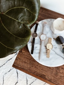 Several wristwatches and a pair of sunglasses are arranged on a round marble tray placed on a wooden table. A large green leaf partially covers the left side of the image, and a white textured rug is visible in the background.