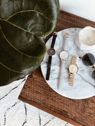 Several wristwatches and a pair of sunglasses are arranged on a round marble tray placed on a wooden table. A large green leaf partially covers the left side of the image, and a white textured rug is visible in the background.