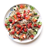 A vibrant salad filled with fresh strawberries, sprouting greens, sliced cucumber, and pieces of ham. The salad is sprinkled with black sesame seeds and served on a white plate.