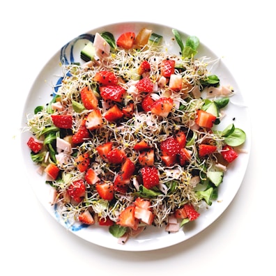 A vibrant salad filled with fresh strawberries, sprouting greens, sliced cucumber, and pieces of ham. The salad is sprinkled with black sesame seeds and served on a white plate.