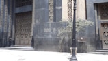 An ornate building facade with intricate metalwork designs on the doors and windows is depicted. The structure features a dark, matte surface with geometric patterns. A black lamppost stands in front, accompanied by a small tree and a stone plaque displaying the words 'Banco de São Paulo'.