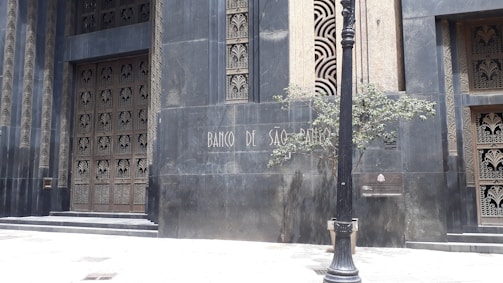 An ornate building facade with intricate metalwork designs on the doors and windows is depicted. The structure features a dark, matte surface with geometric patterns. A black lamppost stands in front, accompanied by a small tree and a stone plaque displaying the words 'Banco de São Paulo'.