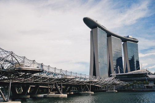 A modern architectural scene featuring a unique building with three towers topped by a structure resembling a ship. In the foreground, there is an intricate, geometric bridge with a lattice-style design over water.