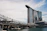 A modern architectural scene featuring a unique building with three towers topped by a structure resembling a ship. In the foreground, there is an intricate, geometric bridge with a lattice-style design over water.