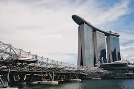 A modern architectural scene featuring a unique building with three towers topped by a structure resembling a ship. In the foreground, there is an intricate, geometric bridge with a lattice-style design over water.