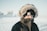A person wearing a thick winter coat with a furry hood stands outside in a snowy landscape. The hat is black and prominently features a logo. The background is a blurred snowy scene with indistinct buildings.
