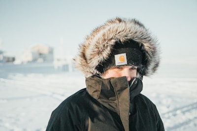 A person wearing a thick winter coat with a furry hood stands outside in a snowy landscape. The hat is black and prominently features a logo. The background is a blurred snowy scene with indistinct buildings.
