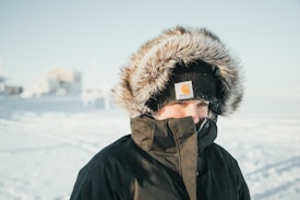 A person wearing a thick winter coat with a furry hood stands outside in a snowy landscape. The hat is black and prominently features a logo. The background is a blurred snowy scene with indistinct buildings.