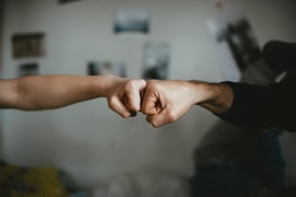 Two fists are bumping together in a gesture of friendship or agreement. The background is slightly blurred, with indistinct images and a soft focus giving a casual and intimate atmosphere.