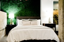 A cozy bedroom features a bed with white linens and gray cushions, flanked by two wooden nightstands with modern lamps. The back wall has a textured green finish resembling a vertical garden, creating a refreshing contrast. Natural elements are highlighted by a large picture on the side depicting a serene landscape.