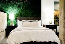 A cozy bedroom features a bed with white linens and gray cushions, flanked by two wooden nightstands with modern lamps. The back wall has a textured green finish resembling a vertical garden, creating a refreshing contrast. Natural elements are highlighted by a large picture on the side depicting a serene landscape.