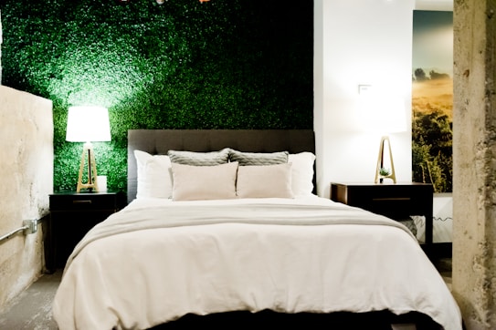 A cozy bedroom features a bed with white linens and gray cushions, flanked by two wooden nightstands with modern lamps. The back wall has a textured green finish resembling a vertical garden, creating a refreshing contrast. Natural elements are highlighted by a large picture on the side depicting a serene landscape.