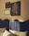 The image features an interior wall adorned with framed artworks above a bed with a textured dark headboard. Three framed pieces are visible: a large painting depicting a stylized tree with white flowers and two cranes, positioned beneath a crescent moon; a smaller frame showcasing botanical illustrations; and another frame with a classic portrait. The room is softly illuminated, enhancing the warm tones of the wall and art.