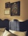 The image features an interior wall adorned with framed artworks above a bed with a textured dark headboard. Three framed pieces are visible: a large painting depicting a stylized tree with white flowers and two cranes, positioned beneath a crescent moon; a smaller frame showcasing botanical illustrations; and another frame with a classic portrait. The room is softly illuminated, enhancing the warm tones of the wall and art.