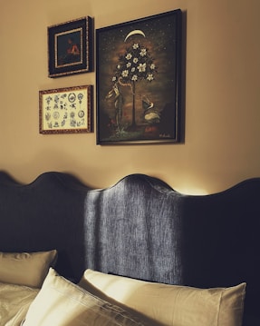 The image features an interior wall adorned with framed artworks above a bed with a textured dark headboard. Three framed pieces are visible: a large painting depicting a stylized tree with white flowers and two cranes, positioned beneath a crescent moon; a smaller frame showcasing botanical illustrations; and another frame with a classic portrait. The room is softly illuminated, enhancing the warm tones of the wall and art.