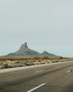 A desert landscape with a prominent rocky peak rising sharply against a pale sky. The foreground features a road stretching into the distance, with desert vegetation lining its sides. The scene conveys a sense of vastness and solitude.