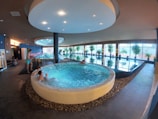 An indoor spa area features a large circular hot tub with people relaxing inside. Adjacent to it is a lap pool lined with windows, allowing natural light to illuminate the space. Potted plants are arranged along the poolside, enhancing the tranquil atmosphere. The ceiling has circular lighting fixtures, and the flooring is tiled. A person is standing near the edge of the hot tub.