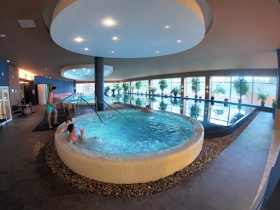 An indoor spa area features a large circular hot tub with people relaxing inside. Adjacent to it is a lap pool lined with windows, allowing natural light to illuminate the space. Potted plants are arranged along the poolside, enhancing the tranquil atmosphere. The ceiling has circular lighting fixtures, and the flooring is tiled. A person is standing near the edge of the hot tub.