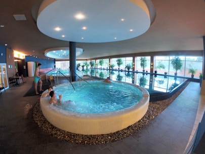 An indoor spa area features a large circular hot tub with people relaxing inside. Adjacent to it is a lap pool lined with windows, allowing natural light to illuminate the space. Potted plants are arranged along the poolside, enhancing the tranquil atmosphere. The ceiling has circular lighting fixtures, and the flooring is tiled. A person is standing near the edge of the hot tub.