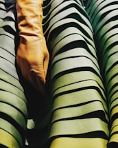 A close-up view of a tan leather glove placed over textured, pleated fabric with various shades of green. The fabric has a layered appearance, creating depth and shadow effects.