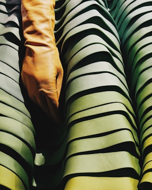 A close-up view of a tan leather glove placed over textured, pleated fabric with various shades of green. The fabric has a layered appearance, creating depth and shadow effects.